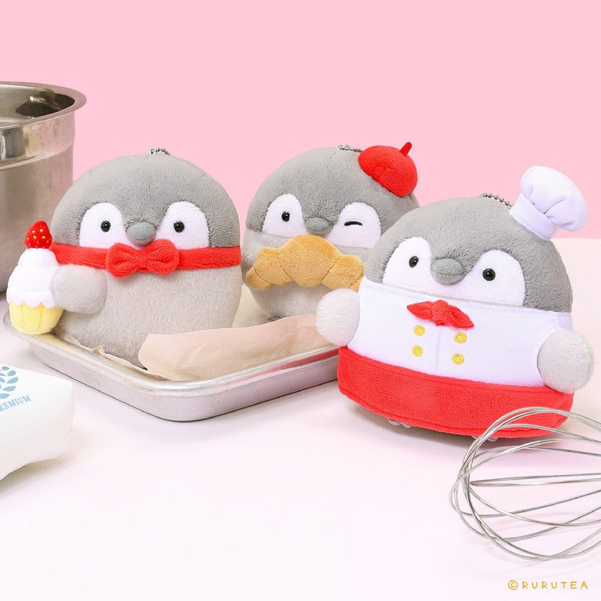 Group of Koupen Chan bakery plush mascot keychains including a penguin with a strawberry cupcake, a penguin eating a croissant, and a chef penguin, arranged with baking tools on a pink background.