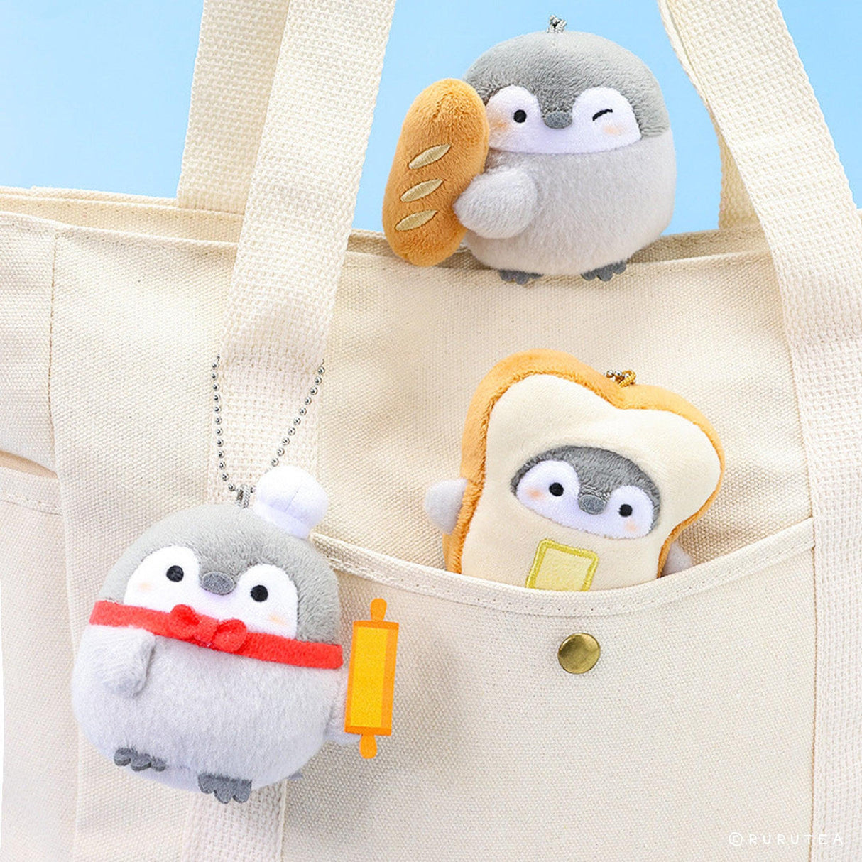 Official Koupen Chan penguin plushie charm keychains featuring Koupen Chan holding a baguette, wearing a toast outfit, and a chef hat with a rolling pin, hanging on a white canvas tote bag
