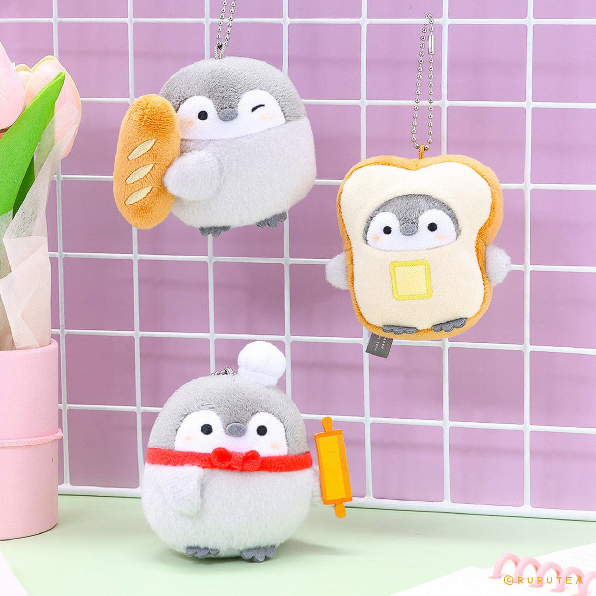 Official Koupen Chan penguin plushie charm keychains featuring Koupen Chan holding a baguette, wearing a toast outfit, and a chef hat with a rolling pin. Bakery theme plush charms hanging on a purple grid wall, cute Japanese penguin accessories for room decoration.