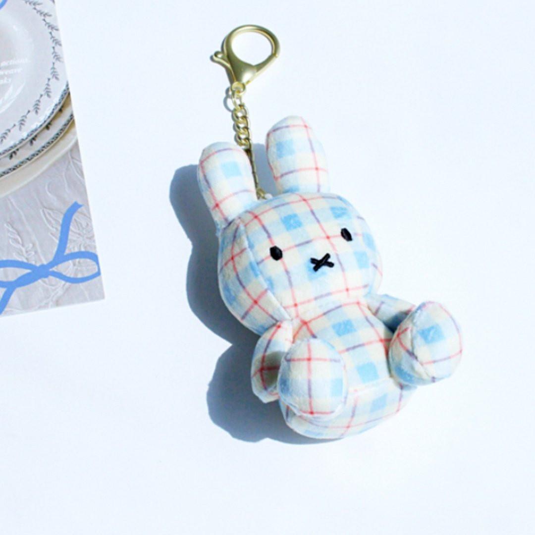 Officially licensed Miffy plush keychain featuring a blue and white plaid/checkered fabric pattern. The soft plush toy is shaped like Miffy, with black embroidered eyes and an 'X' mouth, and includes a gold-tone clasp for use as a bag charm or collector's item.