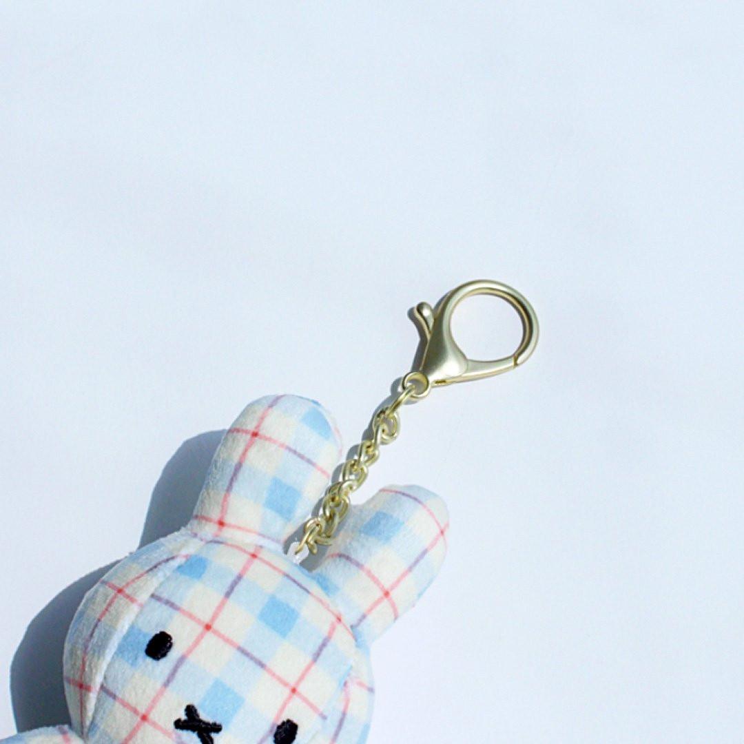 Officially licensed Miffy plush keychain featuring a blue and white plaid/checkered fabric pattern. The soft plush toy is shaped like Miffy, with black embroidered eyes and an 'X' mouth, and includes a gold-tone clasp for use as a bag charm or collector's item.