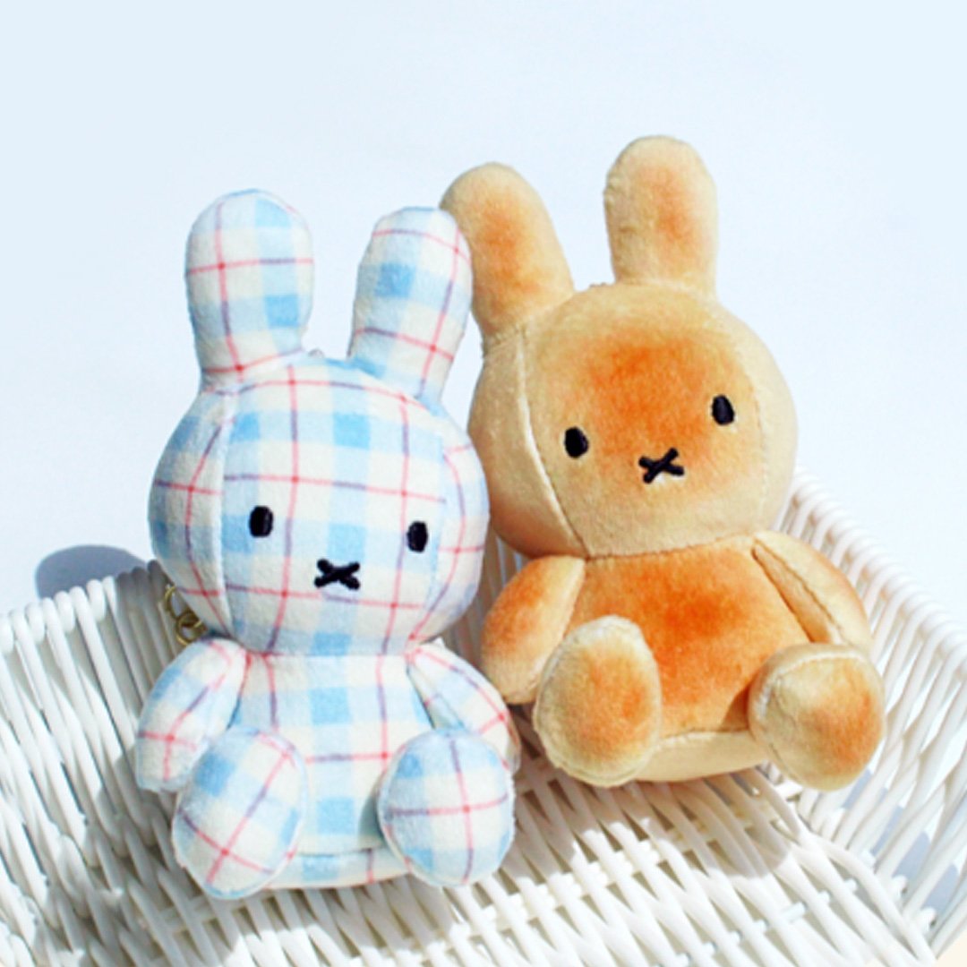 A pair of officially licensed Miffy plush doll keychains seated in a white basket. The set includes two styles: one in bread or toast with its orange-brown color, and one in a light blue, white, and red plaid/checkered fabric. Both Miffy dolls feature embroidered eyes and the signature 'X' mouth.