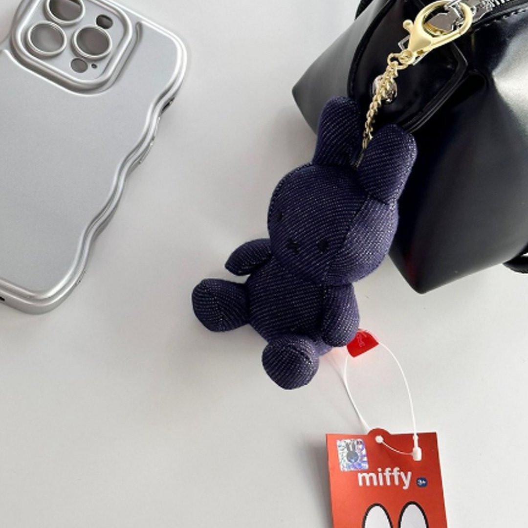 Officially licensed Miffy plush keychain featuring a denim fabric . The soft plush toy keychain attached to a black bag against silver smart phone on the grey table .