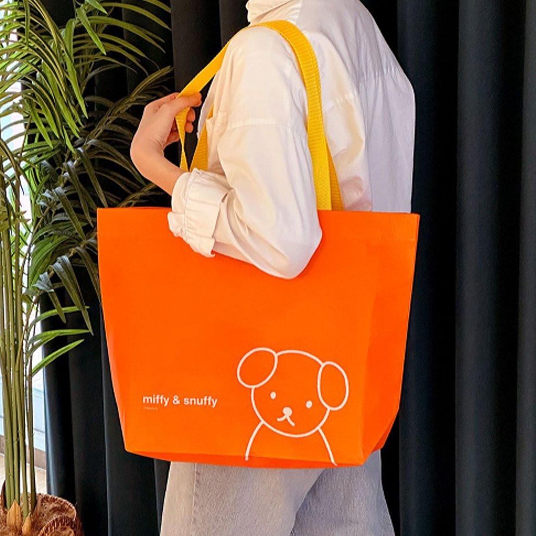 Person holding Officially licensed Miffy & Snuffy Reusable Shopping Bags. Made from durable, wipeable tarpaulin material. Bright orange bag with Miffy and the "miffy & snuffy" text. Excellent for groceries and heavy-duty utility.