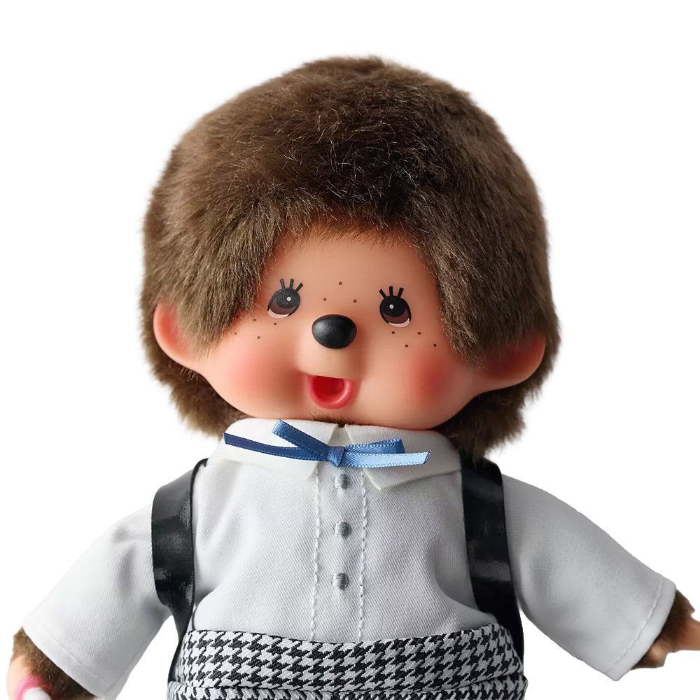 An officially licensed Monchhichi plush doll dressed in a school uniform with houndstooth pants and wearing a black patent leather backpack on its back