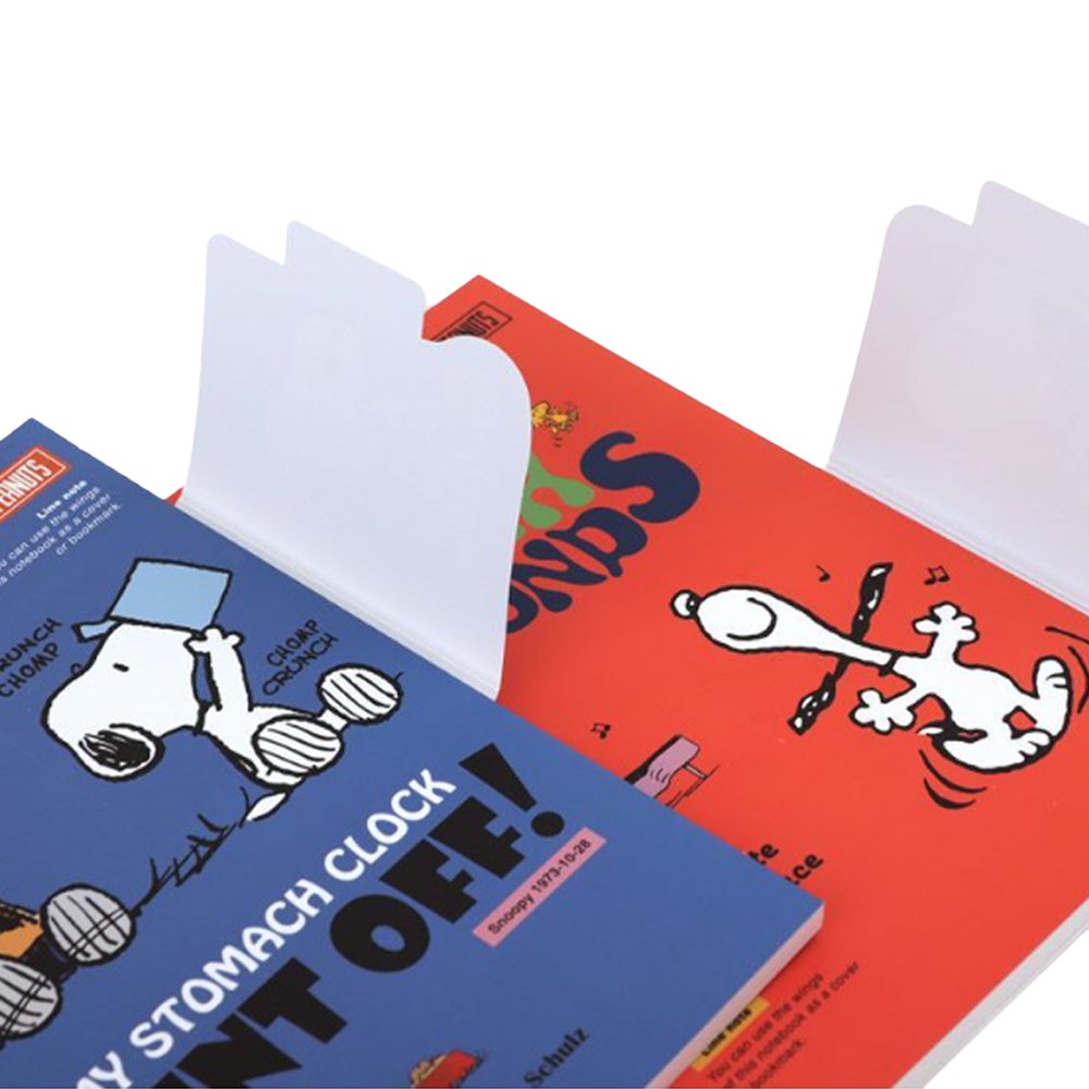 Two officially licensed Peanuts notebooks in red and blue featuring Snoopy and Charlie Brown. The image demonstrates the unique "wing" cover flaps being used as bookmarks and page markers within the lined journal pages.