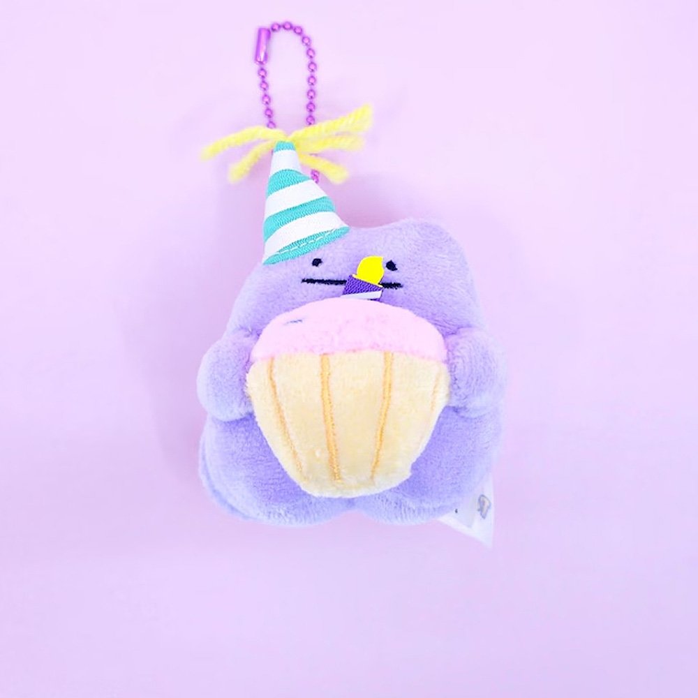 A Pokémon Ditto plush keychain holding a celebration cupcake and wearing a striped party hat with yellow yarn on top, attached to a colorful ball chain