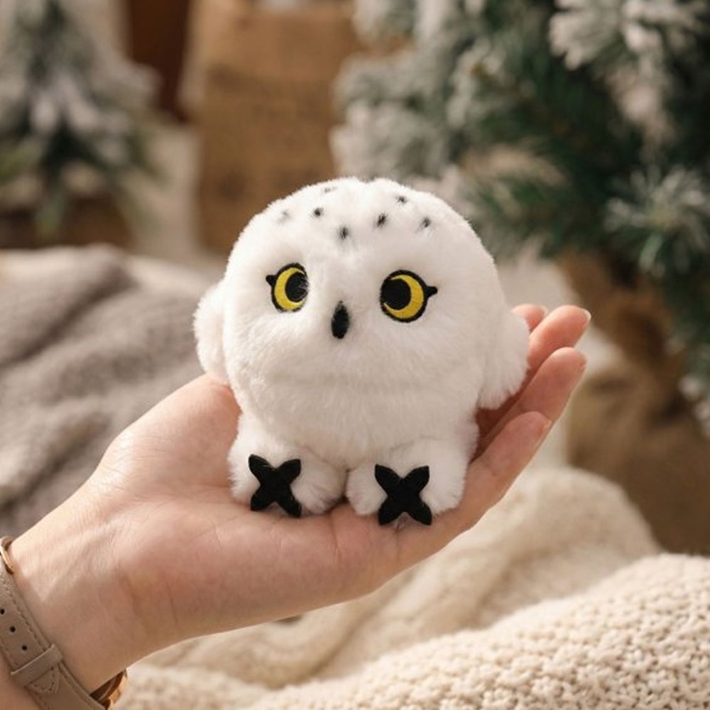 An adorable, round snowy owl plush keychain with fluffy white fur and black speckles on its head and tail. The plush features large, bright yellow embroidered eyes, a tiny black beak, and prominent black talons peeking out from its spherical body.