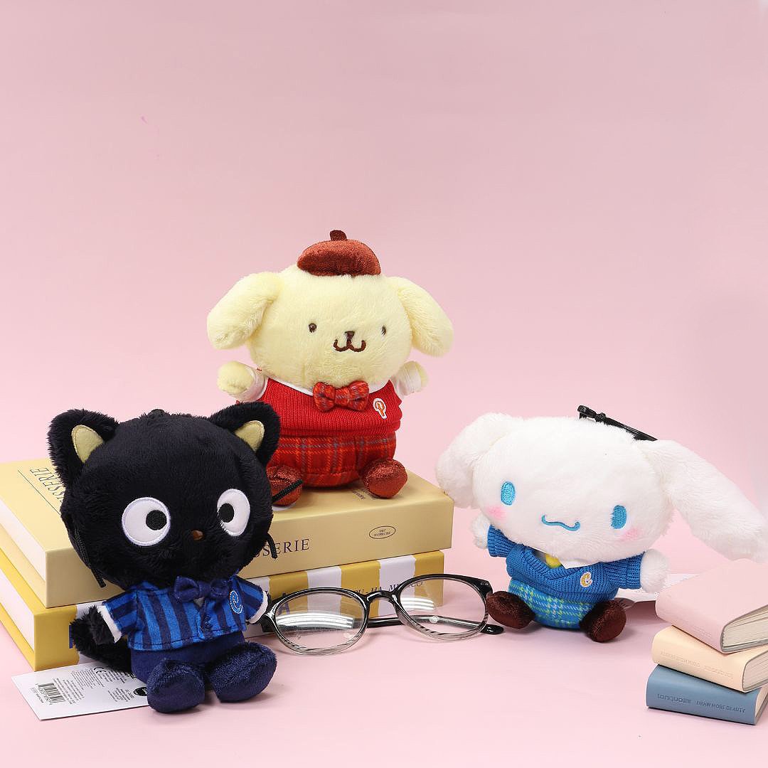 A set of three officially licensed Sanrio plush dolls from the school uniform series: Chococat, Pompompurin, and Cinnamoroll. Chococat wears blue plaid, Pompompurin wears red plaid, and Cinnamoroll wears a blue sweater and plaid skirt. The plush toys are posed on stacked books with a pair of eyeglasses in the foreground, perfect for collectors.
