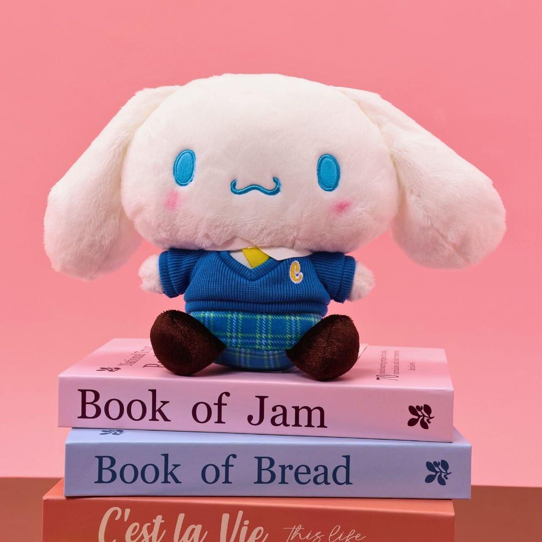 Sanrio officially licensed Cinnamoroll plush doll dressed in a school uniform. He wears a blue plaid skirt, a V-neck blue sweater, a yellow tie, and is seated on a stack of books with colorful spines. The soft, white plush features Cinnamoroll's iconic blue-embroidered eyes and swirling mouth, making it a perfect collectible toy.