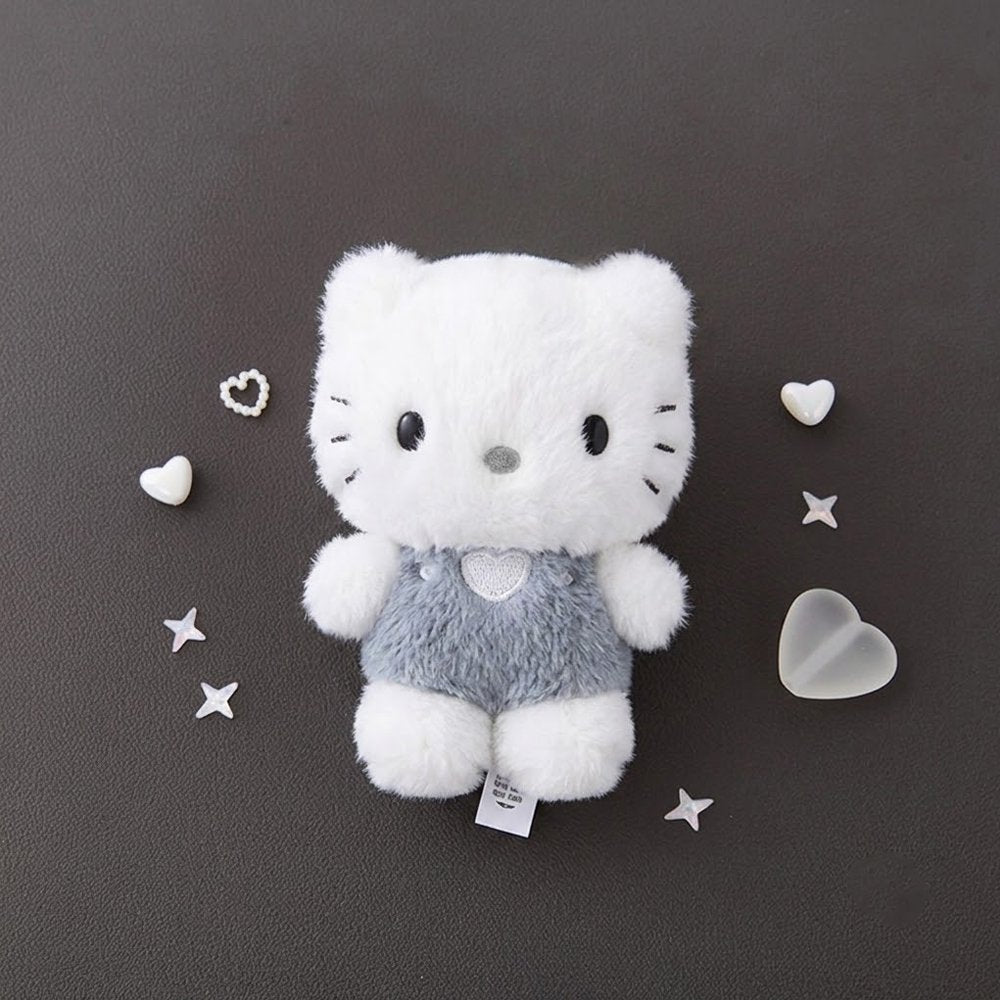 A fluffy white Dear Daniel plush doll keychain wearing textured grey overalls with a heart patch, centered on a dark grey background surrounded by small decorative plastic hearts and stars.