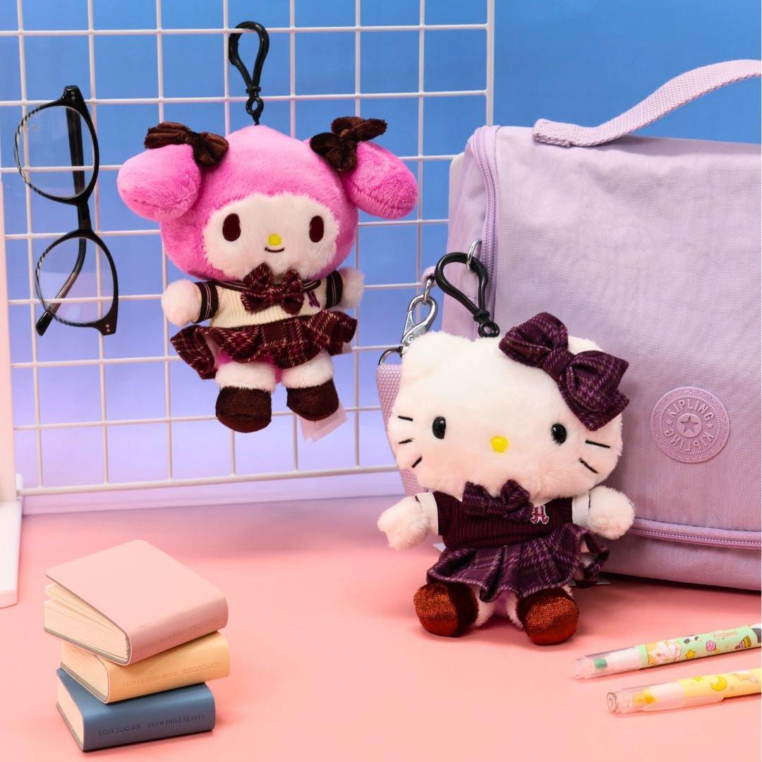 Two small, officially licensed Sanrio plush keychains, Hello Kitty and My Melody, both wearing matching maroon and plaid school uniforms. They are hanging from a white wire grid, with a stack of small books and a light purple bag in the background. Perfect for bag charms or collectors.