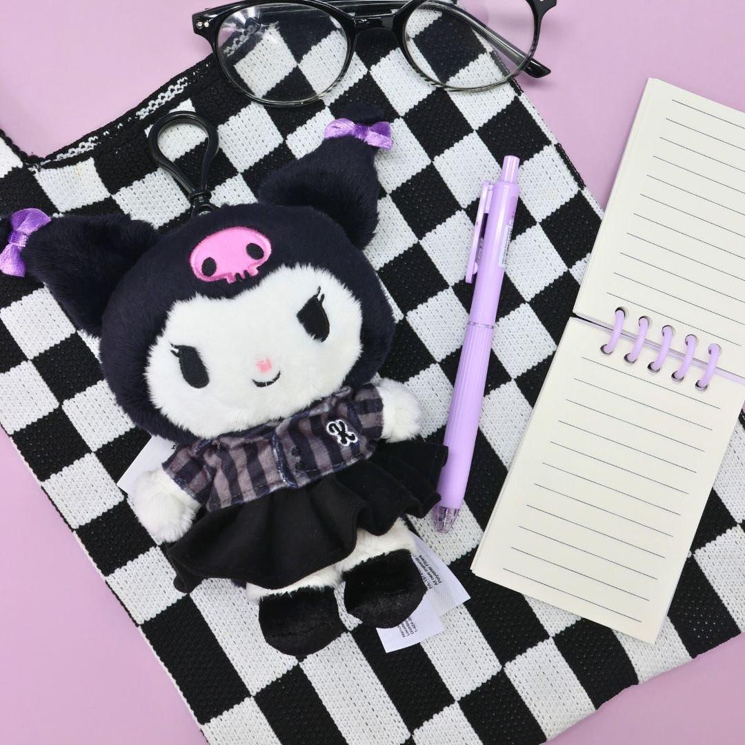 Officially licensed Sanrio Kuromi plush keychain wearing a black and grey plaid school uniform dress with black bows. The keychain is lying on a black and white checkerboard patterned bag or textile, with a lavender pen, notepad, and black glasses nearby. A perfect collectible Kuromi bag charm and accessory.