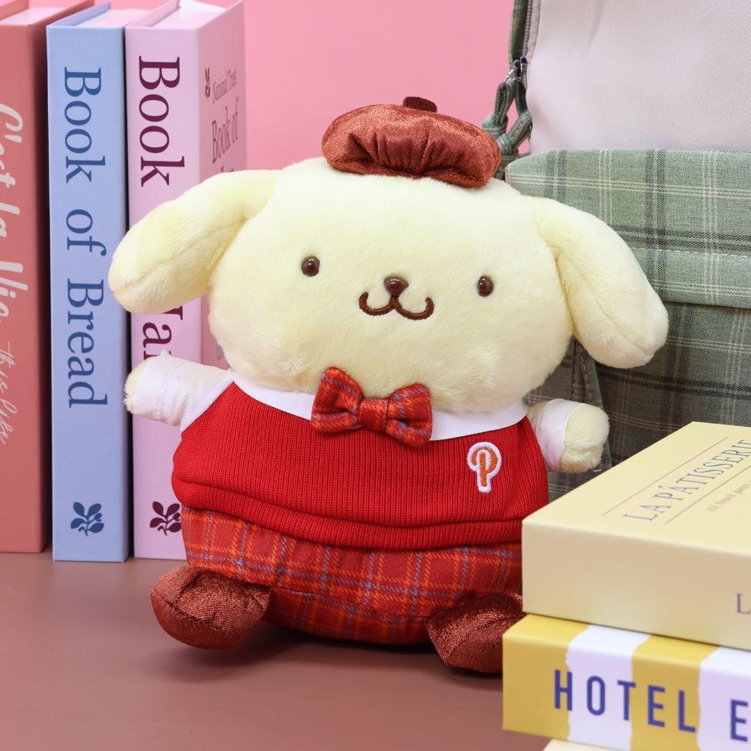 Officially licensed Sanrio Pompompurin plush doll in a collectible school uniform outfit. Pompompurin wears a red knitted sweater vest, a plaid bow tie, matching plaid shorts, and a brown beret hat. The plush is sitting on a desk next to stacked, colorful books, making it a perfect Sanrio collector's item or display piece.