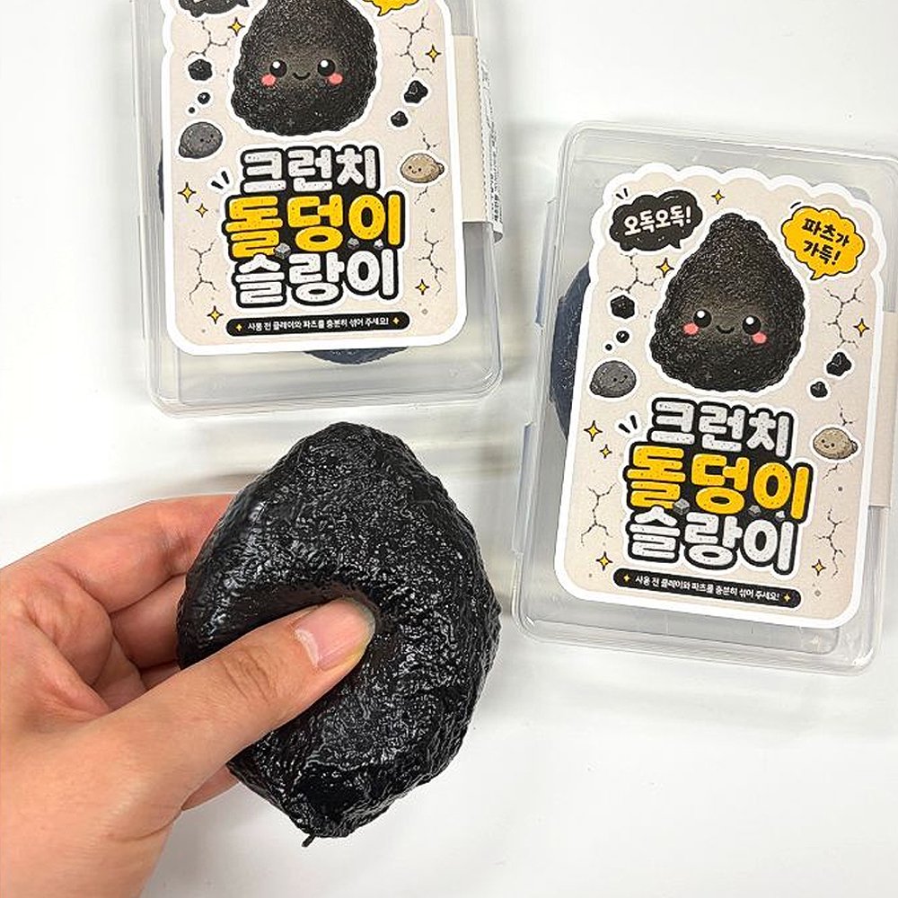 Hand squeezing a dark black stone-shaped squishy slime next to its open clear plastic container and cute cartoon stone sticker packaging, perfect party favor goodie bag filler