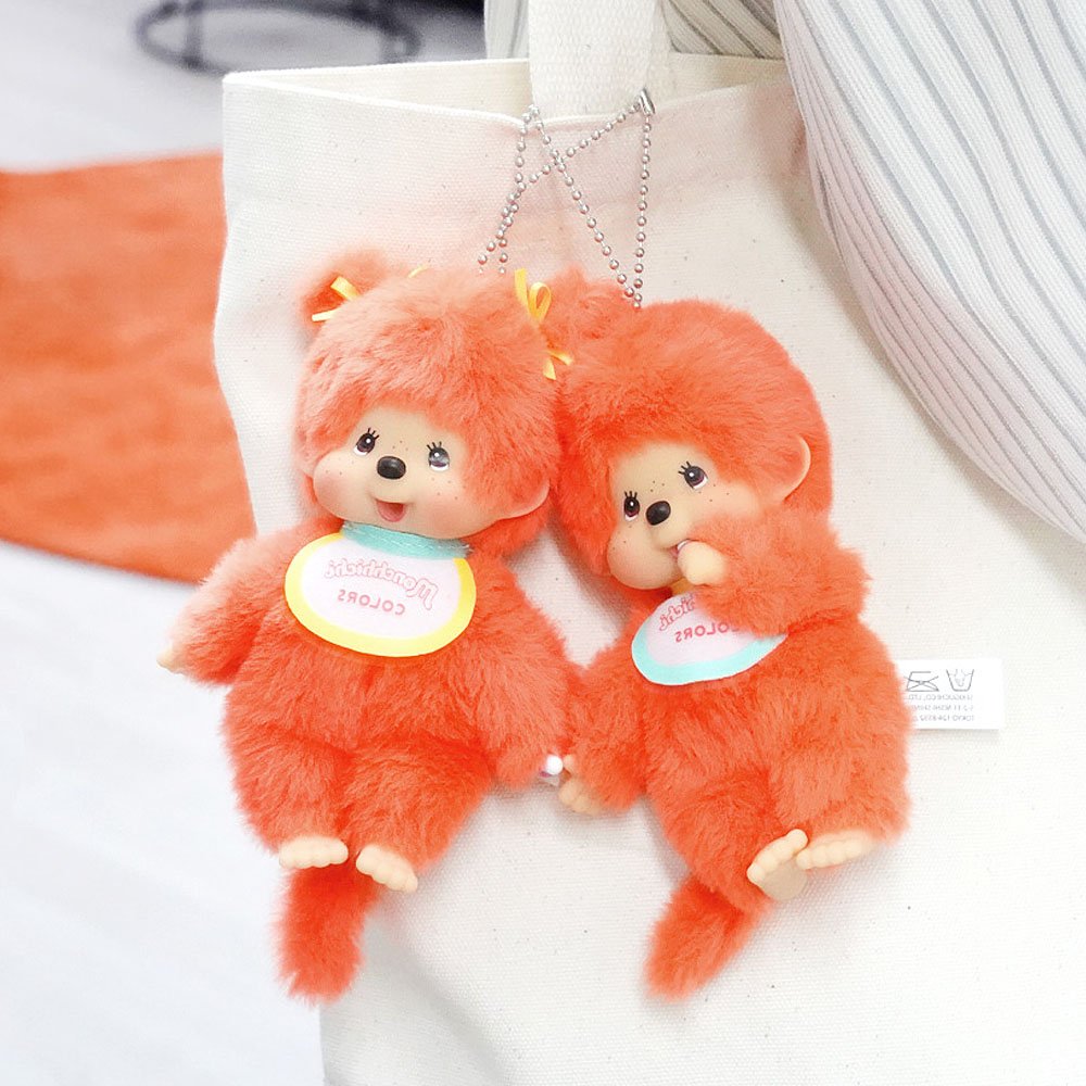 Two orange Monchhichi plush monkey toys with tags on a white surface