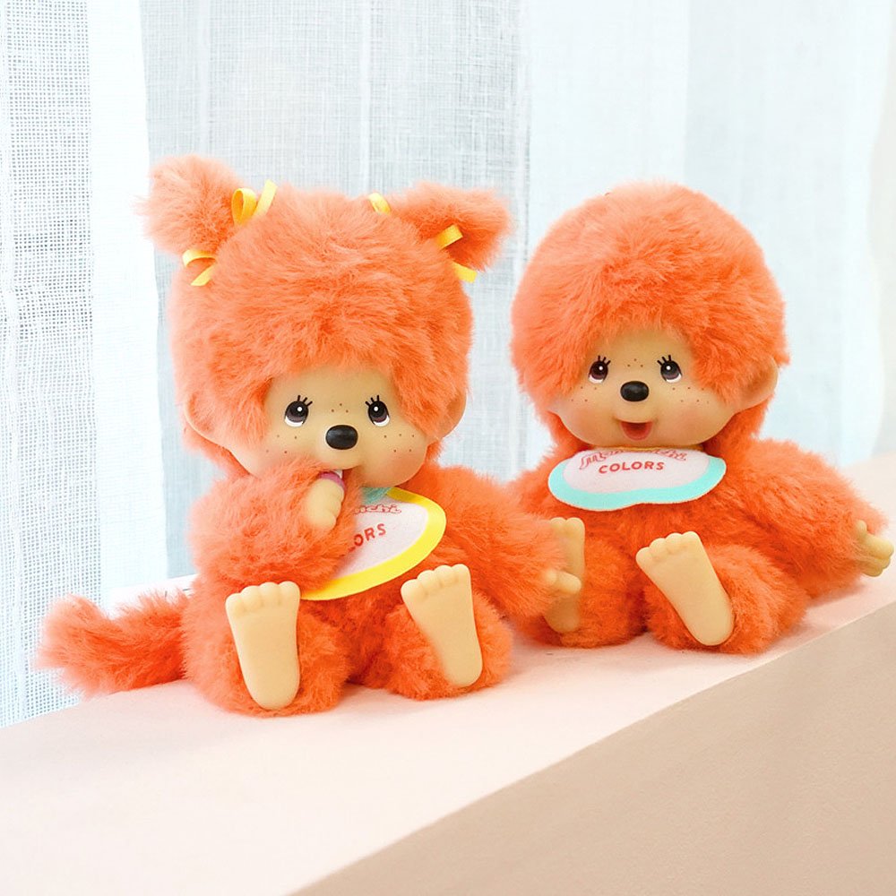 Two orange Monchhichi plush toys with colorful bibs on a light background