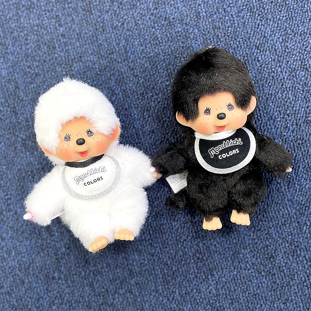 Two plush toys, one white and one black, with 'Monchhichi' branding on a textured blue background.