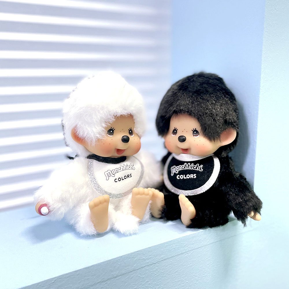 Two Monchhichi dolls, one white and one black, sitting on a light blue surface with a white background.