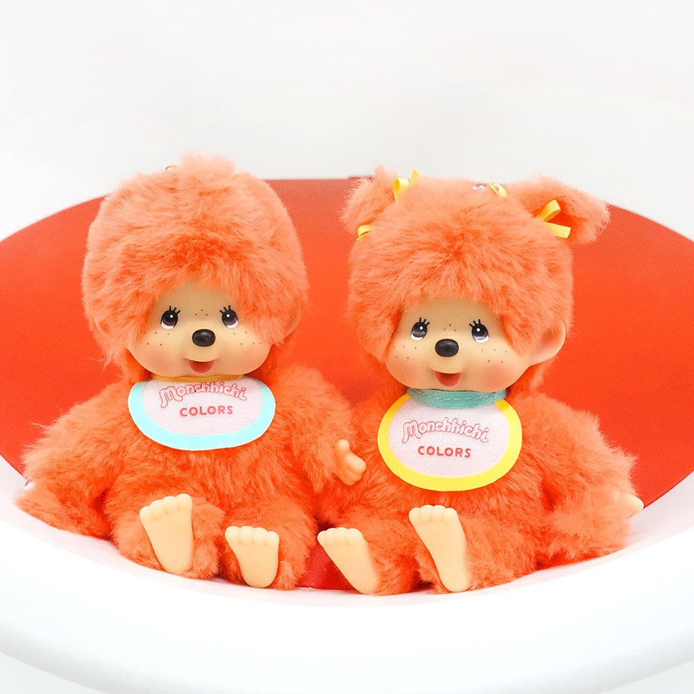 Two orange Monchhichi plush toys with colorful labels on a white background