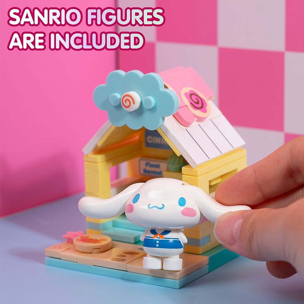 Hand holding a small white Cinnamoroll figure with blue accents in front of a colorful toy house on a pink and white checkered background.