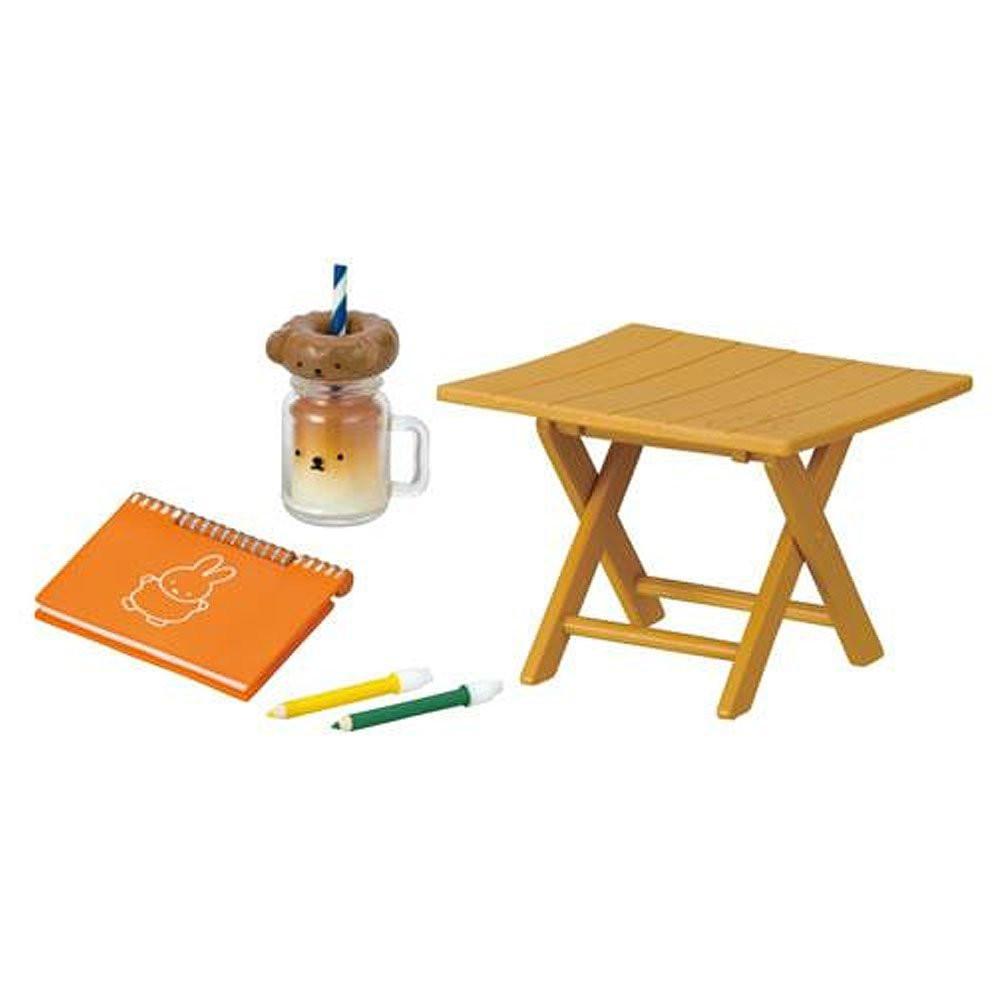 Small wooden table with an orange Miffy notebook, two pens, a glass with a straw, and a small jar on a white background.