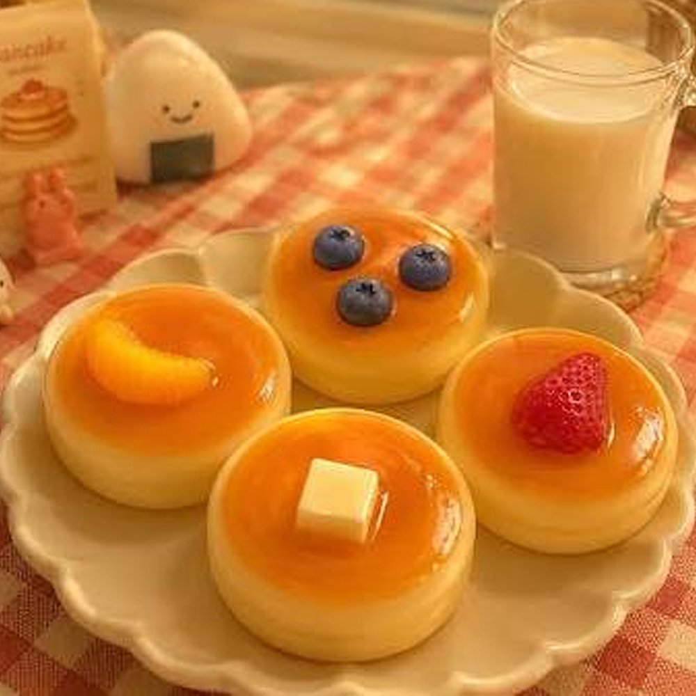 Four realistic pancake squishies with blueberry, strawberry, orange slice, and butter toppings arranged on a plate with a glass of milk on a cozy cafe-style table, perfect non-edible dessert gift idea