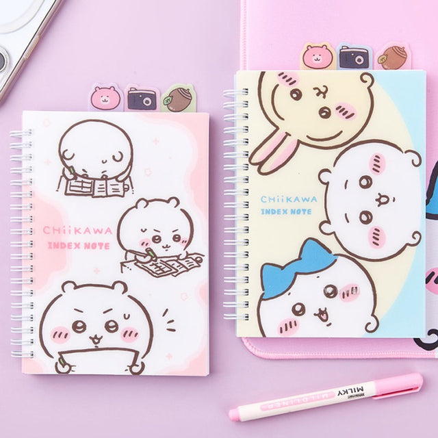 Officially licensed Chiikawa spiral notebooks with index tabs, featuring Chiikawa studying on a pink cover, while the other shows the trio on a yellow and blue background; both include built-in tabs shaped like a bear, a camera, and an acorn.