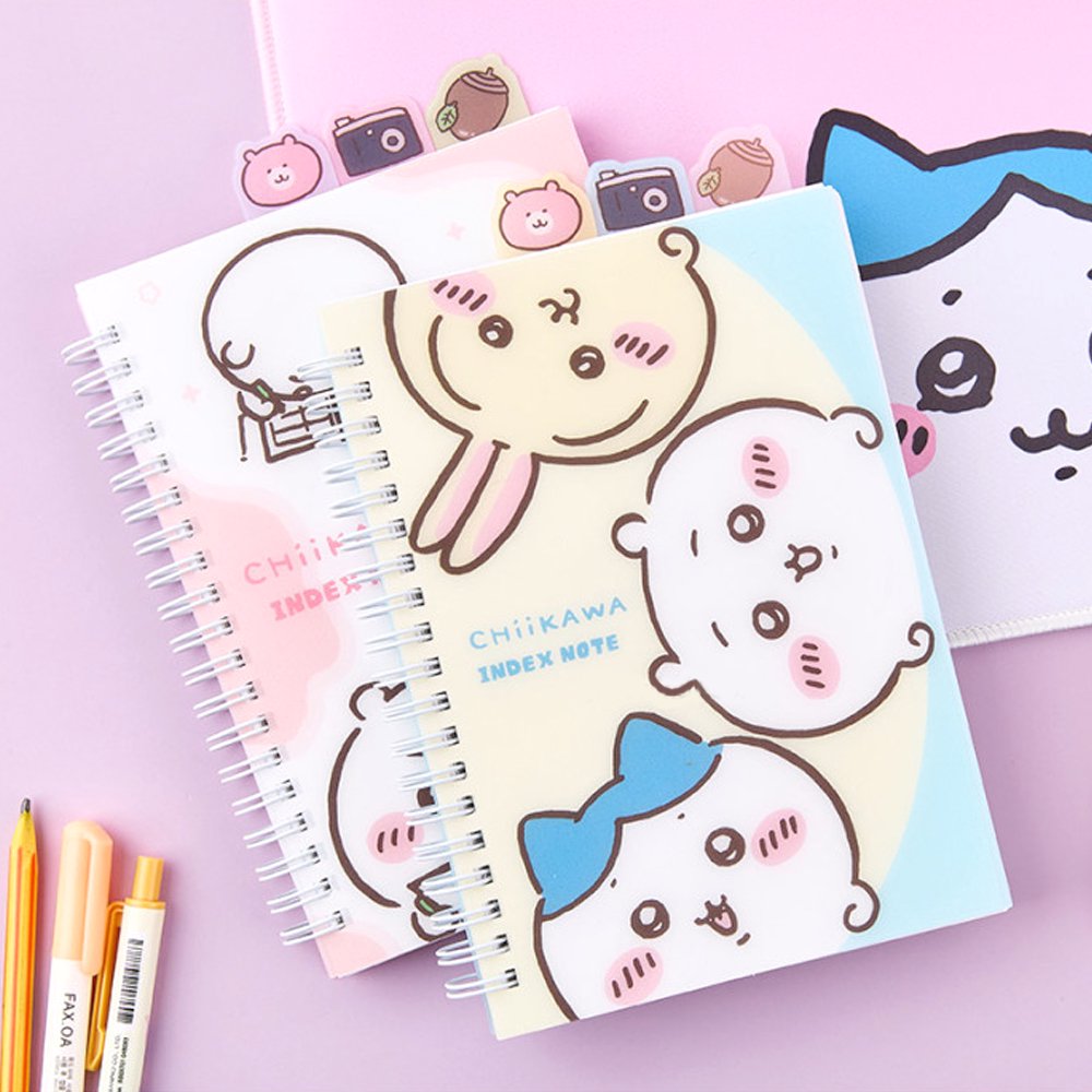 Officially licensed Chiikawa spiral notebooks with index tabs, featuring Chiikawa studying on a pink cover, while the other shows the trio on a yellow and blue background; both include built-in tabs shaped like a bear, a camera, and an acorn.