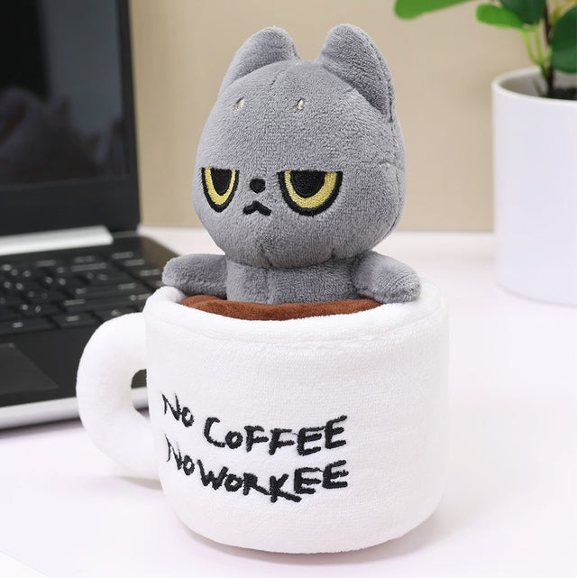 Black cat plush cat toy in a white mug with 'No Coffee No Workee' text, placed on a desk with a laptop and plant in the background.