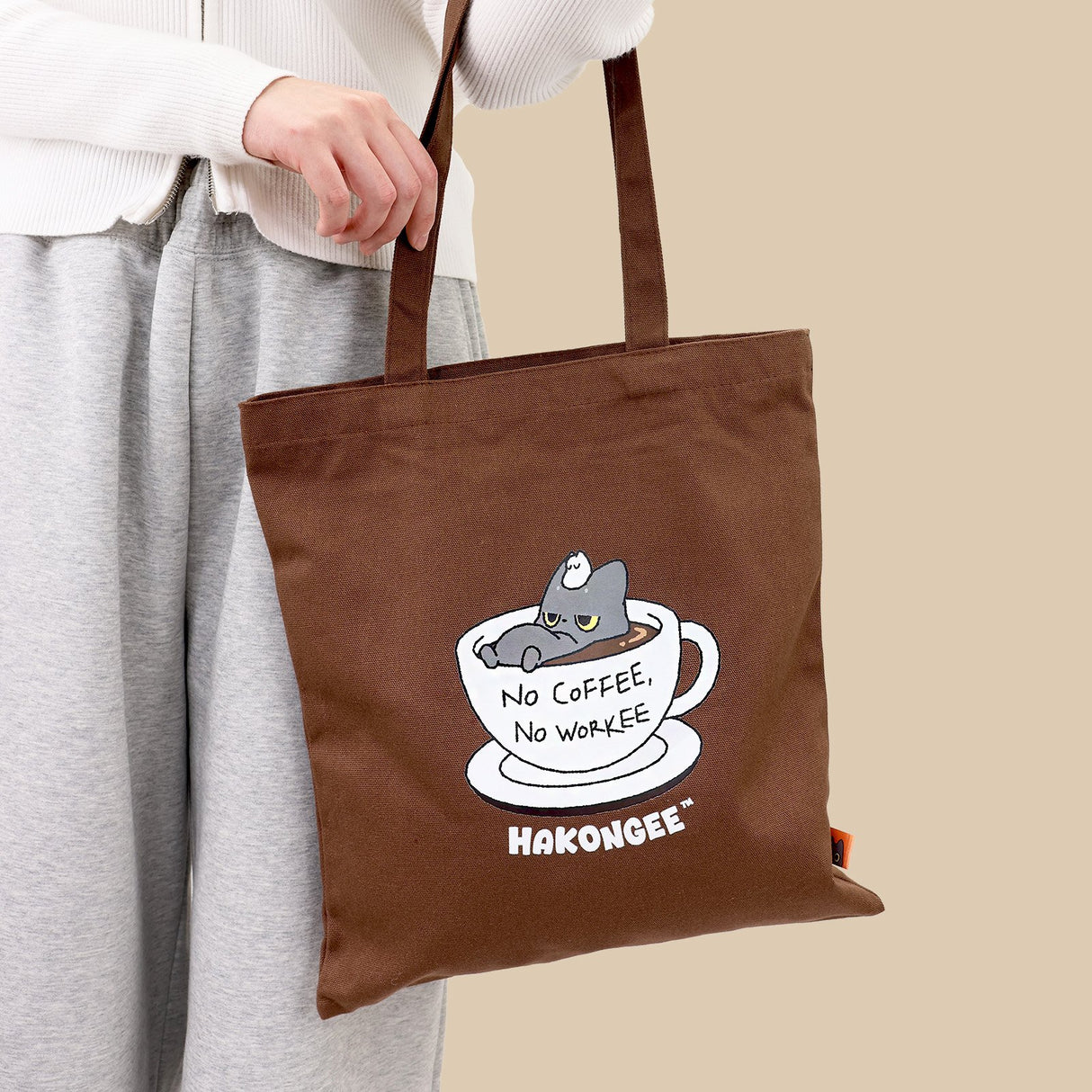 Brown tote bag with a black cat graphic and text held by a person against a beige background