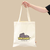Tote bag with 'Hakongee' logo and black cat illustration held by a hand against a beige background