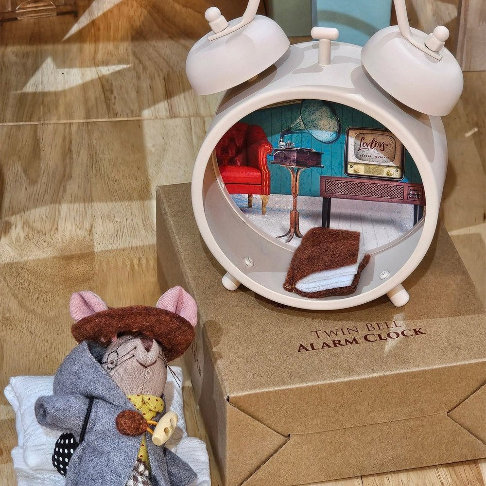 Levlovs' traveling mouse plush wearing a hooded coat and hat stands next to a cream-colored alarm clock structure outfitted as a tiny bedroom