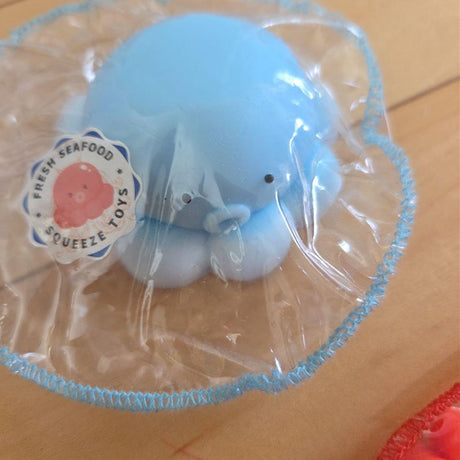 Close-up of the Light Blue Seafood Octopus Mochi Squishy Toy Keychain in a round plastic tray with a Fresh Seafood Squeeze Toys sticker. Features a soft squishy texture, cute black dot eyes, and a small round mouth, making it an aesthetic fidget toy and sensory stress relief charm.