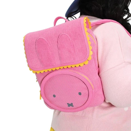A person wearing a pink sweater carries an officially licensed pink corduroy Miffy backpack on their back. The backpack's flap is shaped like rabbit ears and has yellow scalloped trim. A circular window on the front displays Miffy's face.