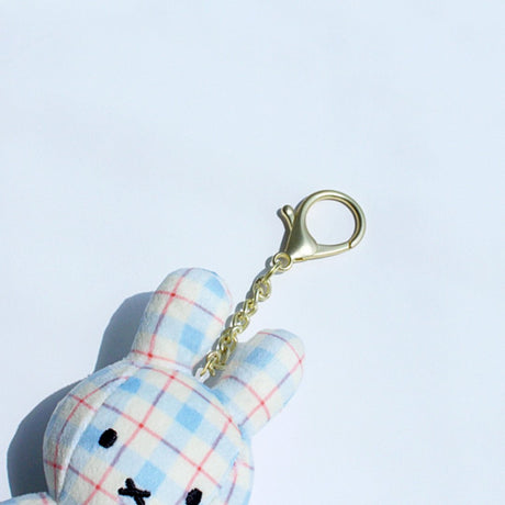 Officially licensed Miffy plush keychain featuring a blue and white plaid/checkered fabric pattern. The soft plush toy is shaped like Miffy, with black embroidered eyes and an 'X' mouth, and includes a gold-tone clasp for use as a bag charm or collector's item.