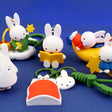 A collection of Miffy dream figure keychains in various poses, including Miffy reading a book, sleeping on a moon, and holding a yellow star, shown with green and blue accessories.