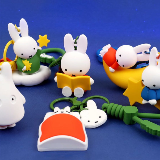 A collection of Miffy dream figure keychains in various poses, including Miffy reading a book, sleeping on a moon, and holding a yellow star, shown with green and blue accessories.