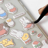 Two officially licensed Miffy Fuzzy Cushion Sticker sheets in cream and mint colors, showcased alongside a decorated phone case and earbud case. The sticker sheets are filled with small, textured, raised stickers depicting Miffy the bunny and her friends engaging in daily activities like baking and gardening.
