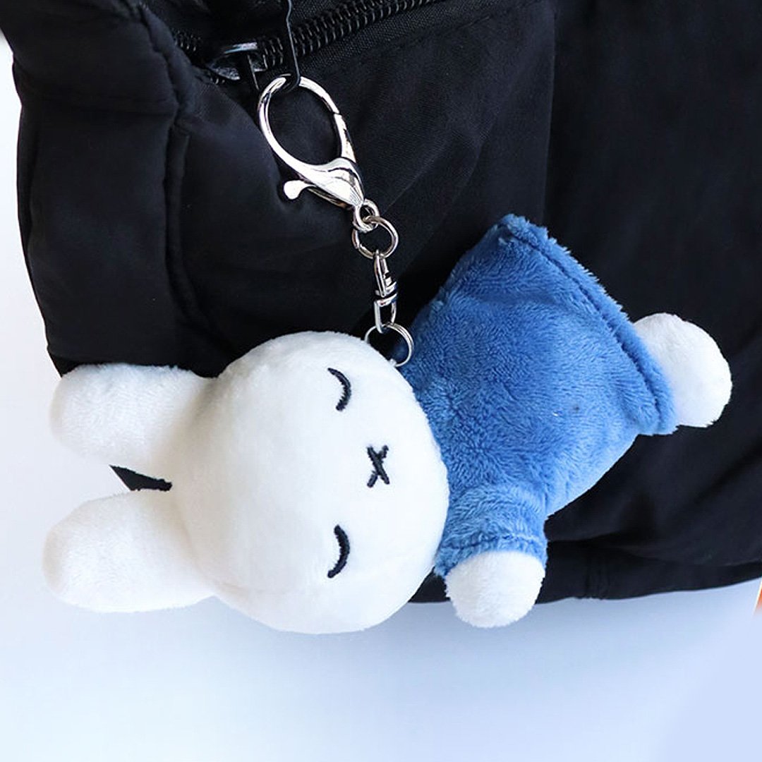 Officially licensed Miffy sleeping plush keychain featuring embroidered closed eyes and an 'X' mouth. Miffy plush wearing green top. The soft plush toy keychain attached to a black  backpack .