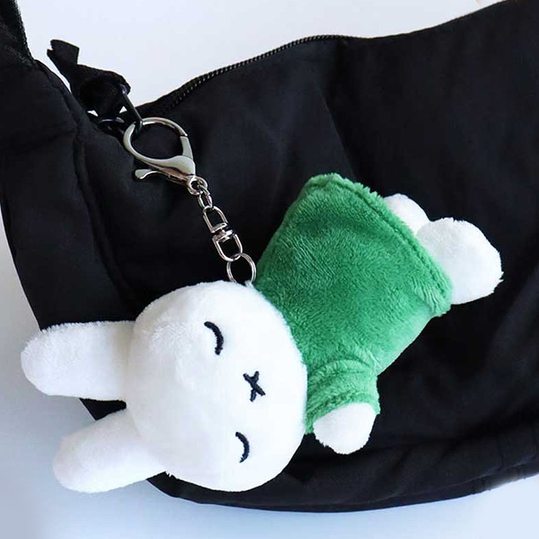 Officially licensed Miffy sleeping plush keychain featuring embroidered closed eyes and an 'X' mouth. Miffy plush wearing green top. The soft plush toy keychain attached to a black  backpack .