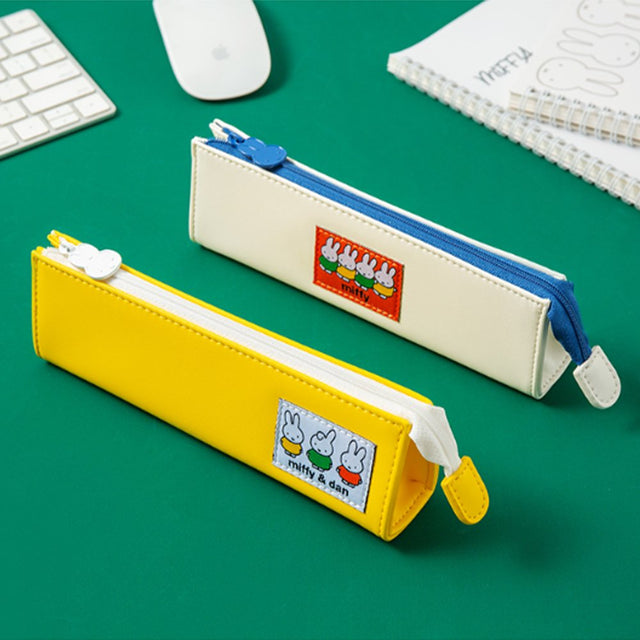 Two officially licensed slim, triangular Miffy pencil cases, featuring one in bright yellow with a white zipper and a "miffy & dan" patch, and the other in cream-colored with a bold blue zipper and a red "miffy" patch.
