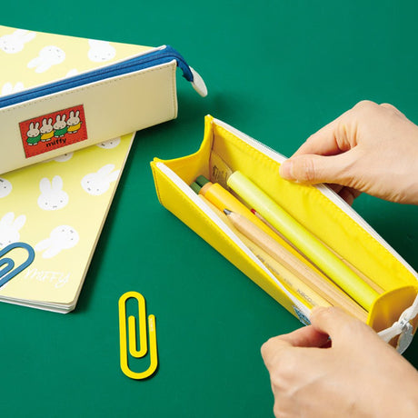Two officially licensed slim, triangular Miffy pencil cases, featuring one in bright yellow with a white zipper and a "miffy & dan" patch, and the other in cream-colored with a bold blue zipper and a red "miffy" patch.

