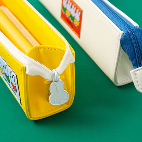 Two officially licensed slim, triangular Miffy pencil cases, featuring one in bright yellow with a white zipper and a "miffy & dan" patch, and the other in cream-colored with a bold blue zipper and a red "miffy" patch.

