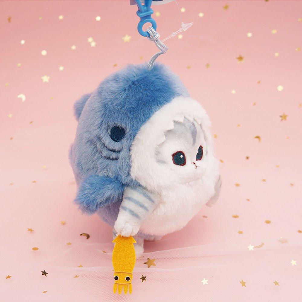 An officially licensed Mofusand shark cat plush keychain featuring a fluffy gray tabby kitten wearing a blue shark onesie with embroidered details