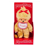 Officially licensed Monchhichi girl plush doll featuring unique golden-beige fur, a long tail, and a cute vinyl face with freckles. The doll has a bright red ribbon tied on its head and wears a white bib with a pink-and-white checkered border that reads "Monchhichi".
