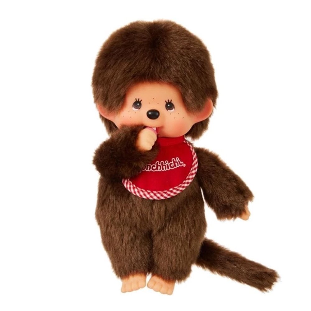 Officially licensed Monchhichi boy plush doll with soft brown fur, a long tail, and an adorable vinyl face with freckles. The doll is wearing a red bib with a checkered border and white text reading "Monchhichi" while holding its hand up to its mouth in its iconic thumb-sucking pose.
