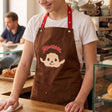An officially licensed brown Monchhichi apron with red adjustable snap-button neck straps and front utility pockets