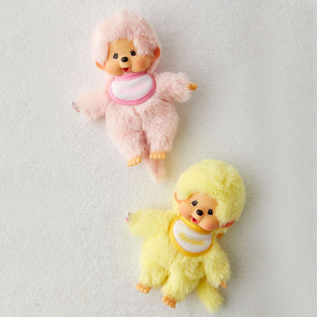 A set of two Monchhichi-style doll plushies. The top doll is pink, and the bottom is yellow, Each plush doll has a tan ABS face, brown eyes, freckles, and is sucking its thumb or finger. They all wear a small bib and have a long, thin tail.