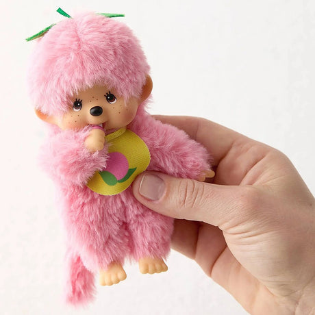A person holding soft and flurry Monchhichi plush doll. officially licensed, peach pink color ,peach -themed Monchhichi plush doll has a tan ABS face, brown eyes, freckles, wear a peach -print bib, and a green stem detail on its head. Monchhichi plush a soft, fluffy texture. Ideal for collection as a charming gift, or for home decor